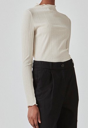 Femme portant un haut crème côtelé à manches longues avec un léger volant au col et aux poignets, associé à un pantalon noir taille haute.