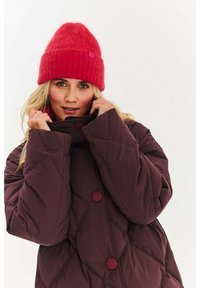 Bordowa pikowana kurtka z dużymi czerwonymi guzikami i wysokim kołnierzem. Noszona z jaskrawo czerwonym prążkowanym dzianinowym czapeczką, z małą metką z logo.