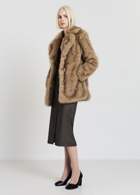 Cappotto in finta pelliccia di colore marrone chiaro con colletto ampio e tasche frontali. Indossato su un abito nero lungo fino al ginocchio e scarpe con tacco alto nere.