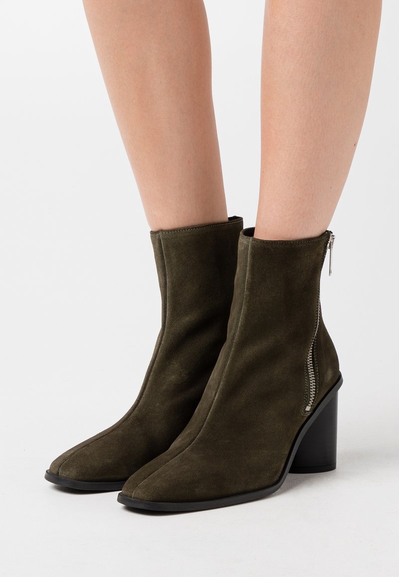 Bottines en daim vert olive avec un bout pointu, une fermeture éclair latérale et un talon épais noir.