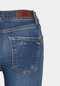 Jeans de mezclilla azul con una etiqueta de cuero marrón que presenta "LTB", costuras naranjas y una textura desgastada en la zona del bolsillo trasero.