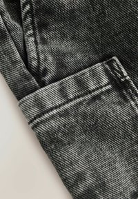 Tejido de mezclilla negra con un acabado texturizado, que presenta un dobladillo doblado. La superficie muestra un patrón gris claro y costuras en contraste.