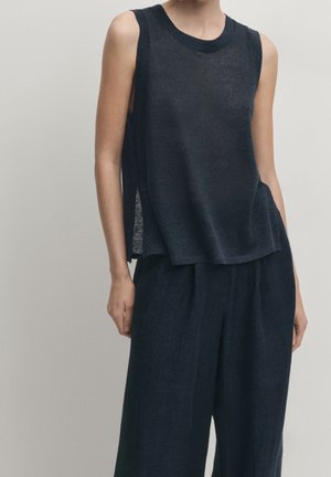Personne portant un haut noir ample sans manches et un pantalon noir à jambes larges, debout devant un fond clair uni.