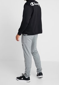 Felpa nera con logo "Champion" bianco sulla schiena, abbinata a pantaloni da jogging grigio chiaro e scarpe da ginnastica nere con accento del logo.