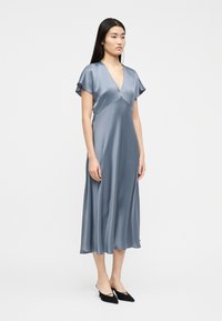 Γυναίκα στέκεται φορώντας ένα λαμπερό, γαλάζιο φόρεμα μήκους μέχρι τον μέσο της γάμπας, με κοντά μανίκια και V-λαιμόκοψη, συνδυασμένο με μαύρα μυτερά παπούτσια με μικρό τακούνι.