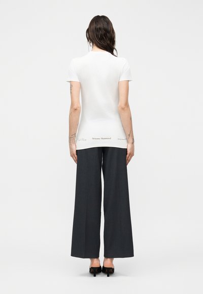 Γυναίκα με μακριά σκούρα μαλλιά, φορώντας λευκό κοντομάνικο εφαρμοστό πουκάμισο και μαύρο παντελόνι με φαρδιά πόδια, όρθια με την πλάτη προς τα εμπρός.