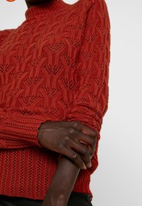 Röd stickad tröja med hög krage, med ett texturerat mönster och ribbade muddar. Materialet verkar mjukt och lätt.