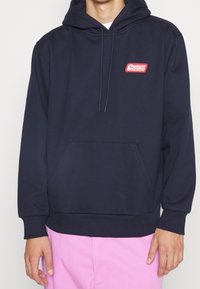 Sudadera azul marino con bolsillo frontal y logo rojo de "Carhartt Services" combinada con pantalones rosa claro.
