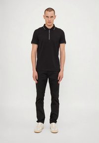 Άνδρας φορώντας μαύρο πόλο μπλούζα με λευκό κείμενο στο περιθώριο, μαύρο παντελόνι και λευκά αθλητικά παπούτσια, που στέκεται μπροστά από έναν ουδέτερο λευκό φόντο.