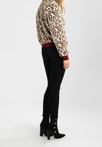 Žena v leopardím vzoru bundě, černých skinny kalhotách a černých kotníkových botách na podpatku se zlatými knoflíky, stojící bokem.