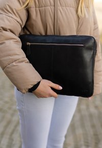 Černý kožený obal na laptop s texturovaným povrchem, vybavený horním zipem a zadní kapsou. Drží ho osoba ve béžové péřové bundě.