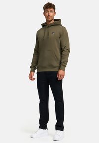 Sudadera verde olivo con bolsillo tipo canguro, capucha ajustable con cordón y un logo negro. Combinada con pantalones negros y zapatillas blancas.