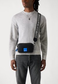 Geantă crossbody neagră cu un accent albastru, închidere cu fermoar și detalii cu logo. Curea ajustabilă cu text, asociată cu un pulover gri cu model cu nervuri.