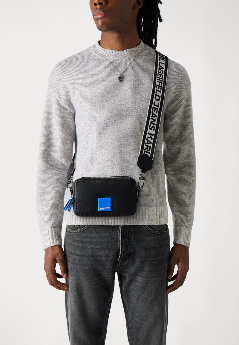Geantă crossbody neagră cu un accent albastru, închidere cu fermoar și detalii cu logo. Curea ajustabilă cu text, asociată cu un pulover gri cu model cu nervuri.