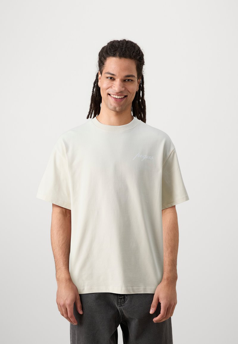 Ljust beige bomull t-shirt med rundad hals, korta ärmar och en liten, broderad logotyp i vitt på vänster sida.