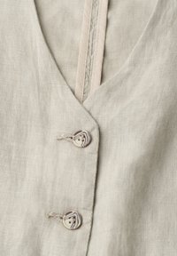 Vêtement en lin beige en gros plan montrant des boutons beige et marron marbrés avec des boutonnières cousues le long de l'ouverture en V.