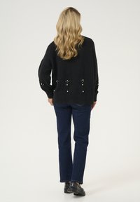 Svart stickad tröja med texturerad detaljering och pärlaccent, i kombination med mörkblå jeans och svarta skor, sedd bakifrån.