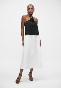 Femme portant un haut halter noir avec un détail floral, associé à une jupe midi blanche et des sandales à talons hauts noires à lanières.
