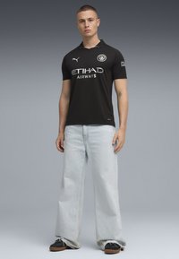 Svart Puma fotbollströja med krage, med en vit logotyp och texten "ETIHAD AIRWAYS". Matchas med ljusblå vida jeans och svarta skor.