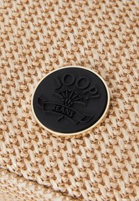 Materiale intrecciato beige con una superficie texturizzata presenta un logo rotondo nero e oro impresso con la scritta "JOOP! JEANS" e una bandiera sottostante.