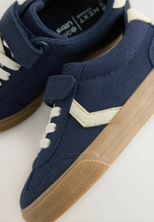 Sneakers blu navy con lacci bianchi e una fascia in velcro, caratterizzati da una striscia laterale beige e una suola in gomma naturale, visualizzati da un'angolazione.