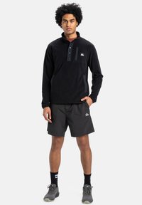 Zwarte fleece pullover met een kwart-rits, voorzien van een borstzak en textuuraccenten, gecombineerd met zwarte shorts en grijze wandelschoenen.