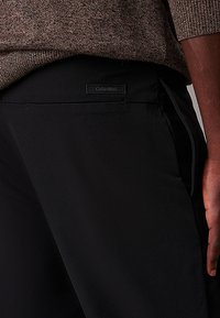 Pantaloni neri Calvin Klein con una texture liscia, vestibilità su misura e un'etichetta del marchio visibile sulla tasca posteriore.