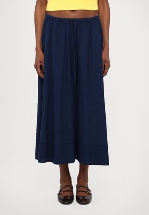 Personne portant une jupe large bleu marine avec une taille à cordon, associée à des chaussures Mary Jane noires et un top court jaune.
