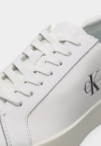 Λευκό αθλητικό παπούτσι Calvin Klein με λευκά κορδόνια, λείο δερμάτινο επάνω μέρος και μαύρο λογότυπο "CK" στο πλάι κοντά στη σόλα.