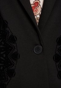 Blazer negro con patrones de encaje texturizado en los laterales, cierre con un solo botón negro y camisa con estampado floral visible en el escote.