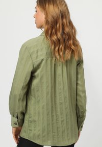 Camisa de manga larga color verde oliva con rayas verticales, fabricada en un tejido suave, con puños abotonados y un ajuste relajado, vista desde la parte de atrás.