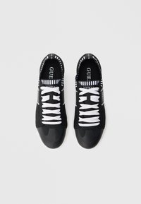 Ζευγάρι μαύρα και λευκά αθλητικά παπούτσια Guess με λευκά κορδόνια και ριγωτό εσωτερικό, σε λευκό φόντο από ψηλά.