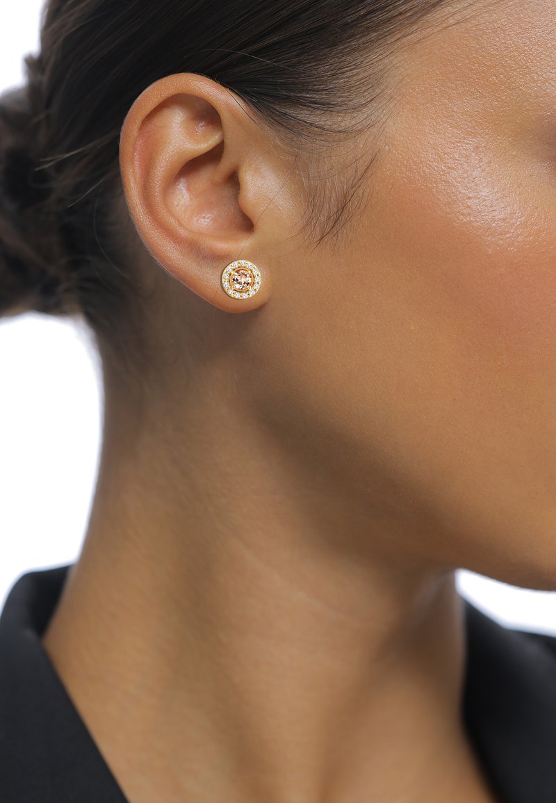 Boucles d'oreilles en métal doré en forme de clou avec une pierre précieuse ronde au centre, entourée de petites pierres transparentes, le tout dans un design circulaire.