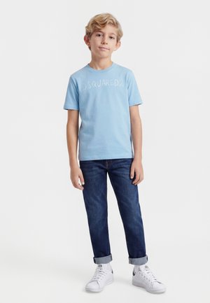 Szőke fiú áll, világoskék DSQUARED2 pólót, sötét felhajtott farmert, fehér tornacipőt és fehér zoknit visel, fehér háttér előtt.