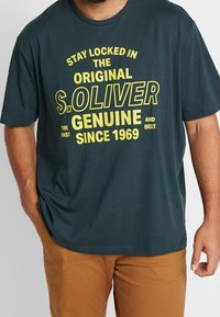 Teal katoenen t-shirt met gele bedrukte tekst, met "S. OLIVER", ronde halslijn en korte mouwen. Gecombineerd met bruine broek.