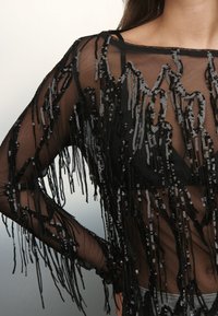 Black sheer top with long fringe made of black and silver sequins, featuring a boat neckline and long sleeves.