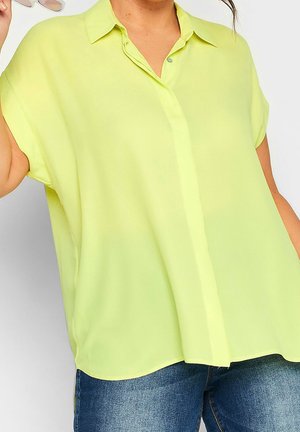 Woman wearing a loose, short-sleeve, button-up lime green blouse paired with blue denim jeans, holding sunglasses in one hand.