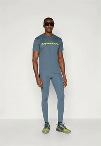 Férfi sportöltözet, amely tartalmaz egy kék rövid ujjú pólót neon logóval, hozzá illő kék leggingset és színes futócipőt.