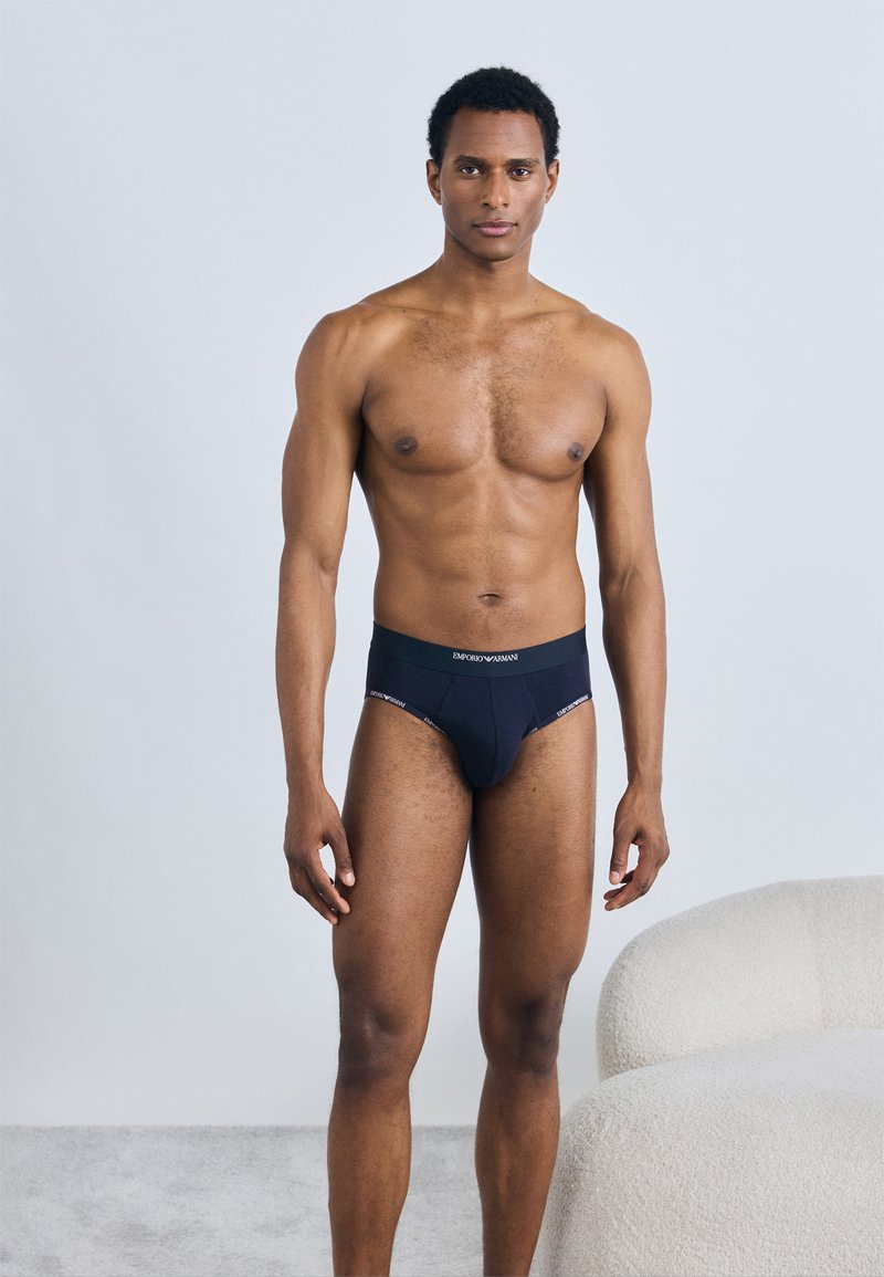 Melni apakšbiksītes ar logotipa jostu, izgatavotas no mīksta auduma. Modelis stāv uz gaismas paklāja blakus krēmkrāsas izliektam dīvānam.