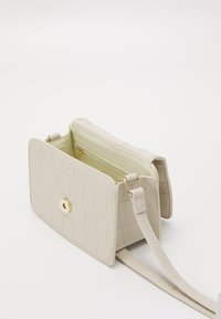 Beżowa torebka ze wzorem krokodyla, z złotym zapięciem na zatrzask, regulowanym paskiem i zielonym wnętrzem, które zawiera wewnętrzną kieszeń na zamek.