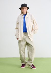 Ein cremefarbenes gestreiftes Hemd, ein blauer T-Shirt, weite hellgraue Hosen, lila-weiße Sneakers und ein schwarzer Bucket Hat. Vor einem grünen Boden.