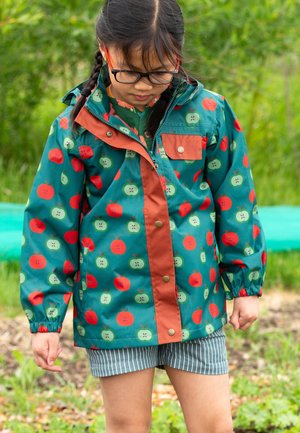 Kind mit geflochtenem Haar und Brille, trägt eine türkise Jacke mit rotem und grünem Apfeldruck, steht im Freien und schaut nach unten.
