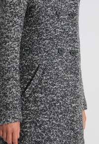 Abrigo texturizado en gris y negro con diseño de doble botonadura, que presenta botones negros y un bolsillo lateral. Material suave y tejido.