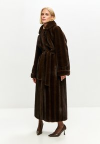 Brun faux fur frakke med en bred krave, bælte i taljen og lange ærmer, der har en vertikal ribbet tekstur og en gulvlængde.