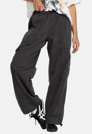 Person trägt lockere dunkle Cargohosen, schwarz-weiße Sneakers und ein weißes T-Shirt mit Aufdruck, steht mit einem Bein über das andere geschlagen.