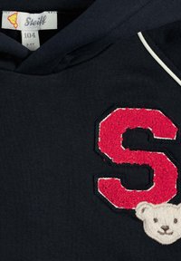 Felpa navy con un grande ricamo rosso a forma di "S" e una toppa di orsetto bianco, con cuciture chiare in contrasto lungo i bordi.