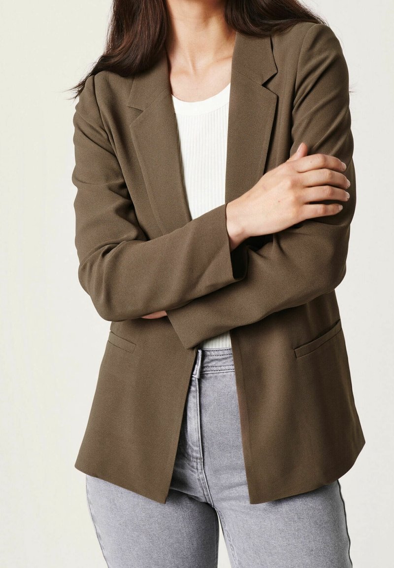 Femme portant un blazer olive, un haut blanc côtelé et un jean gris clair, les bras croisés sur la poitrine devant un fond uni.