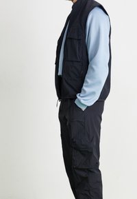 Laivastonsininen hihaton liivi vaaleansinisen pitkähihaisen paidan päällä, jossa on useita taskuja ja joustavat hihansuut, yhdistetty vastaaviin housuihin.