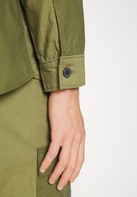 Camicia verde oliva con un design strutturato. Il polsino abbottonato presenta un bottone nero e una tasca applicata di lato. Materiale in cotone con una texture liscia.
