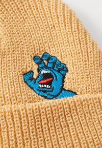 Brede gebreide muts in lichtgeel, met een blauwe geborduurde handmotief met een schreeuwende mond en rode tong, prominent op de voorkant geplaatst.
